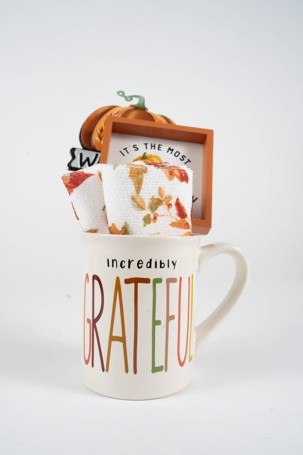 A white ceramic mug with the word "Grateful" in colorful letters and "Incredibly" above in black letters. This Grateful Mug Gift Basket with Free Food Certificate includes a rolled-up festive towel adorned with autumn leaf patterns, a small decorative frame, a mini pumpkin ornament, and a Free Food Certificate.