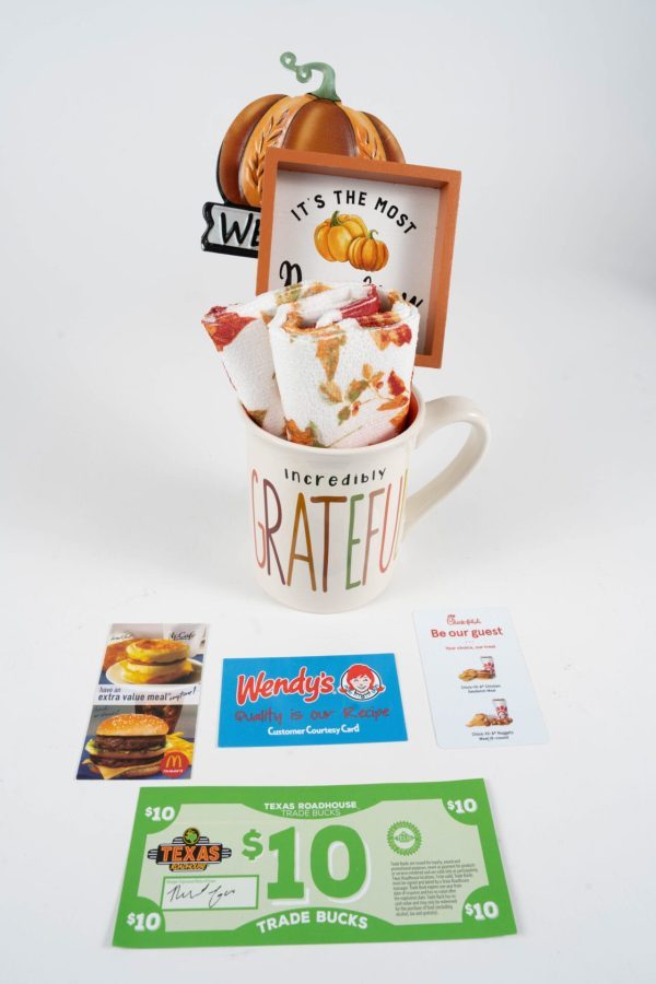 A Grateful Mug Gift Basket with Free Food Certificate, labeled "Incredibly Grateful," contains colorful autumn-themed kitchen towels. Behind it, signs display "It's the Most Wonderful Time of the Year" with a pumpkin image. Arranged below are free food certificates for McDonald's, Wendy's, and Texas Roadhouse.