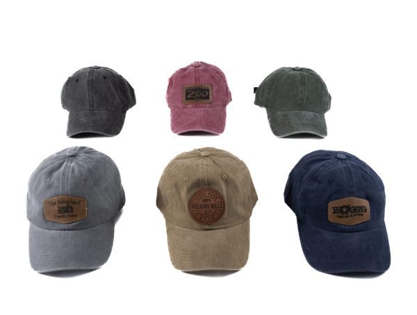 A set of six Custom Hats - Distressed with Leather Patch, showcased in various colors and designs. The top row includes dark gray, maroon, and green caps, while the bottom row highlights light gray, beige with a circular leather patch, and navy blue caps with a rectangular patch.