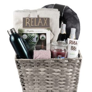 The His & Hers Spa Gift Basket - Large includes a grey wicker basket containing a black water bottle, pink towel, rolled white towel, box labeled "Relax," sparkling beverage bottle, jar candle, Bulldog skincare products, and a grey and black blanket—the ultimate spa gift for both him and her.