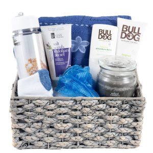 The His & Hers Spa Gift Basket - Medium includes a medium woven basket filled with a silver water bottle, a tube of salt scrub exfoliant, a blue towel with a floral design, two tubes of Bulldog skincare products, a "sandalwood amber" jar candle, and a blue shower pouf—ideal for creating the perfect spa experience for both him and her.