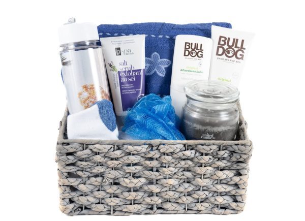 The His & Hers Spa Gift Basket - Medium includes a medium woven basket filled with a silver water bottle, a tube of salt scrub exfoliant, a blue towel with a floral design, two tubes of Bulldog skincare products, a "sandalwood amber" jar candle, and a blue shower pouf—ideal for creating the perfect spa experience for both him and her.