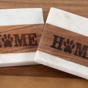 The Pair of Pet Square Coasters - Home, featuring marble coasters with wooden inlays, sit on a wooden surface. Each inlay showcases the word "HOME" with a paw print replacing the letter "O," creating a rustic and pet-friendly home decor aesthetic.