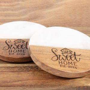 A pair of "Home Sweet Home" round coasters, featuring a half-wood, half-marble design, are placed on a wooden surface. They are engraved with the phrase "Home Sweet Home" accompanied by a small house icon above the text and "Est. 2024" underneath.