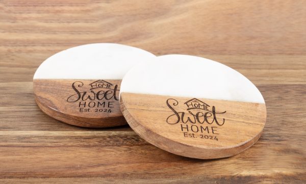 A pair of "Home Sweet Home" round coasters, featuring a half-wood, half-marble design, are placed on a wooden surface. They are engraved with the phrase "Home Sweet Home" accompanied by a small house icon above the text and "Est. 2024" underneath.