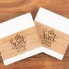 The pair of square coasters, named "Home Sweet Home," showcase a wooden design and feature the phrase "Home Sweet Home Est. 2024." Set against a wooden background, they evoke a warm, homey atmosphere.