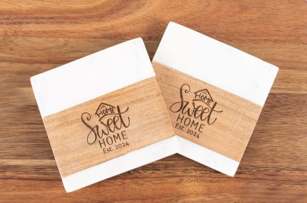 The pair of square coasters, named "Home Sweet Home," showcase a wooden design and feature the phrase "Home Sweet Home Est. 2024." Set against a wooden background, they evoke a warm, homey atmosphere.