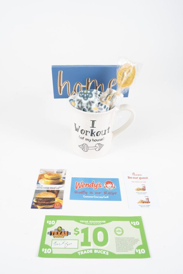 The "I Work Out of the House" Mug Gift Basket with Free Food Certificate is a charming collection that includes a decorative "home" sign, a lollipop, a white "Work Out of the House" mug, a Wendy's gift card, a Texas Roadhouse $10 trade buck, and a Panera Bread free food certificate. The items are neatly arranged against a plain white background.