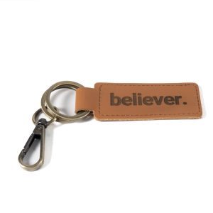 The "Key Chain - Believer" features a sophisticated brown leather tag embossed with the word "believer." in black letters. The tag is securely attached to a metal key ring and clip, all beautifully showcased against a simple white backdrop.