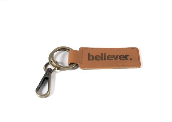 The "Key Chain - Believer" features a sophisticated brown leather tag embossed with the word "believer." in black letters. The tag is securely attached to a metal key ring and clip, all beautifully showcased against a simple white backdrop.