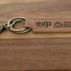 A Custom Key Chain with a metal clasp and two key rings rests on a wooden surface. The leather tag, laser engraved on both sides, features the logos of "eXp Realty" and "Ohio Housing Experts".