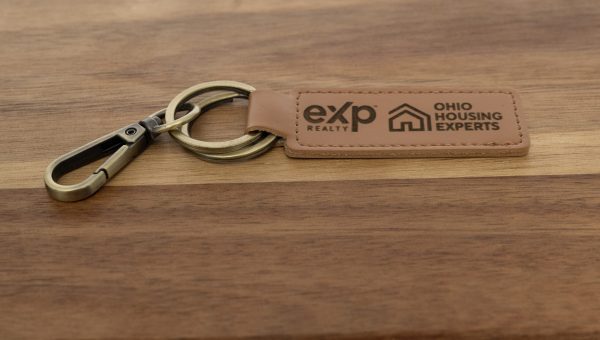 A Custom Key Chain with a metal clasp and two key rings rests on a wooden surface. The leather tag, laser engraved on both sides, features the logos of "eXp Realty" and "Ohio Housing Experts".