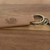 A Custom Key Chain - Laser engraved on one or both sides with the words "Home Sweet Home" lies on a wooden surface. The keychain, made from light brown leather, features a decorative font for the text and includes a metal keyring along with an attached clip.