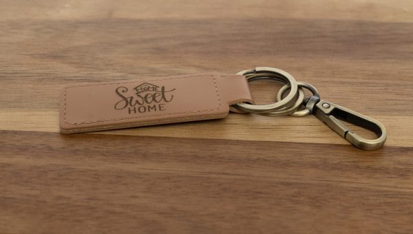 A Custom Key Chain - Laser engraved on one or both sides with the words "Home Sweet Home" lies on a wooden surface. The keychain, made from light brown leather, features a decorative font for the text and includes a metal keyring along with an attached clip.