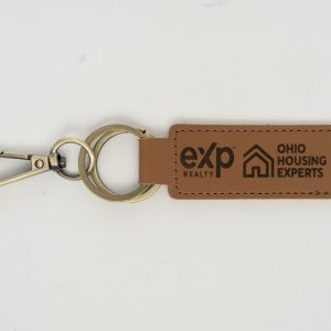 Introducing the Custom Key Chain - Laser engraved on one or Both Sides, crafted from high-quality brown leather and equipped with a sturdy metal keyring and clip. This elegant keychain features professionally laser-engraved logos along with the text "eXp Realty" and "Ohio Housing Experts" on both sides of the strap. The keyring connects seamlessly to both the strap and the clip, creating a cohesive, stylish accessory against a plain white background.