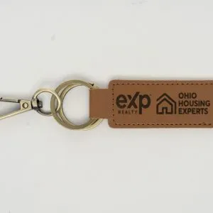 Introducing the Custom Key Chain - Laser engraved on one or Both Sides, crafted from high-quality brown leather and equipped with a sturdy metal keyring and clip. This elegant keychain features professionally laser-engraved logos along with the text "eXp Realty" and "Ohio Housing Experts" on both sides of the strap. The keyring connects seamlessly to both the strap and the clip, creating a cohesive, stylish accessory against a plain white background.