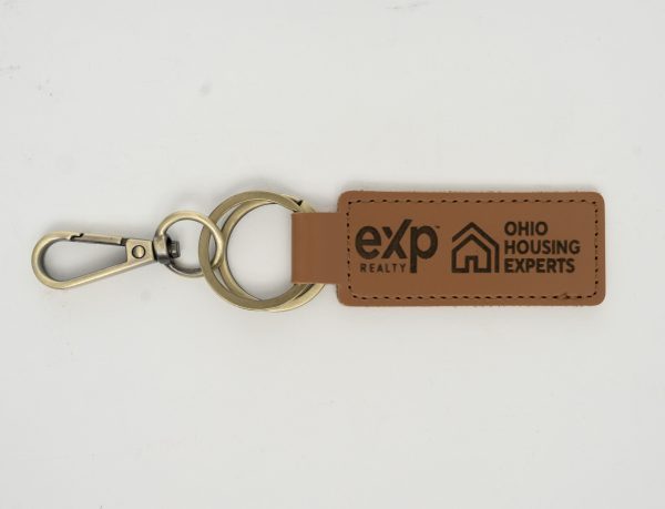 Introducing the Custom Key Chain - Laser engraved on one or Both Sides, crafted from high-quality brown leather and equipped with a sturdy metal keyring and clip. This elegant keychain features professionally laser-engraved logos along with the text "eXp Realty" and "Ohio Housing Experts" on both sides of the strap. The keyring connects seamlessly to both the strap and the clip, creating a cohesive, stylish accessory against a plain white background.