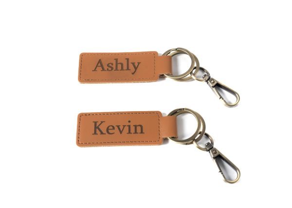 Two keychains from the "Home Sweet Home Key Chains - Set of 2" are shown with rectangular leather tags. The top keychain's tag reads "Ashly" while the bottom keychain's tag reads "Kevin." Each leather tag features attached metal rings and clasps, making them perfect for adding a touch of personalized charm to your home.