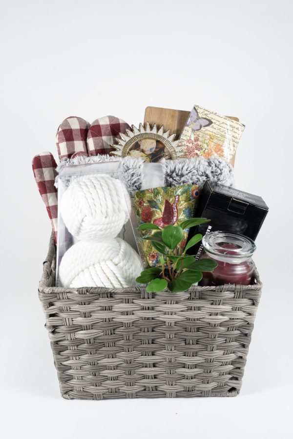 An Adult Gambling Night Basket filled with various items: red and white checkered oven mitts, two rolls of chunky white yarn, a small green potted plant, a red candle in a glass jar, a floral-patterned notebook, and other small household and decorative items perfect for an adult's relaxing night at home.