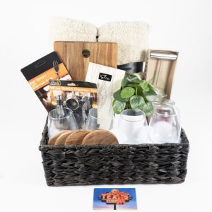 A black woven basket from our Personalized Kitchen & Home Baskets collection contains various items including a custom cutting board, four custom wine glasses, four custom coasters, $75 in Texas Roadhouse certificates, a bottle opener set, two rolled-up white towels, a small plant, and a cheese slicer. A colorful flyer with the text "Thank You" is placed in front of the basket.