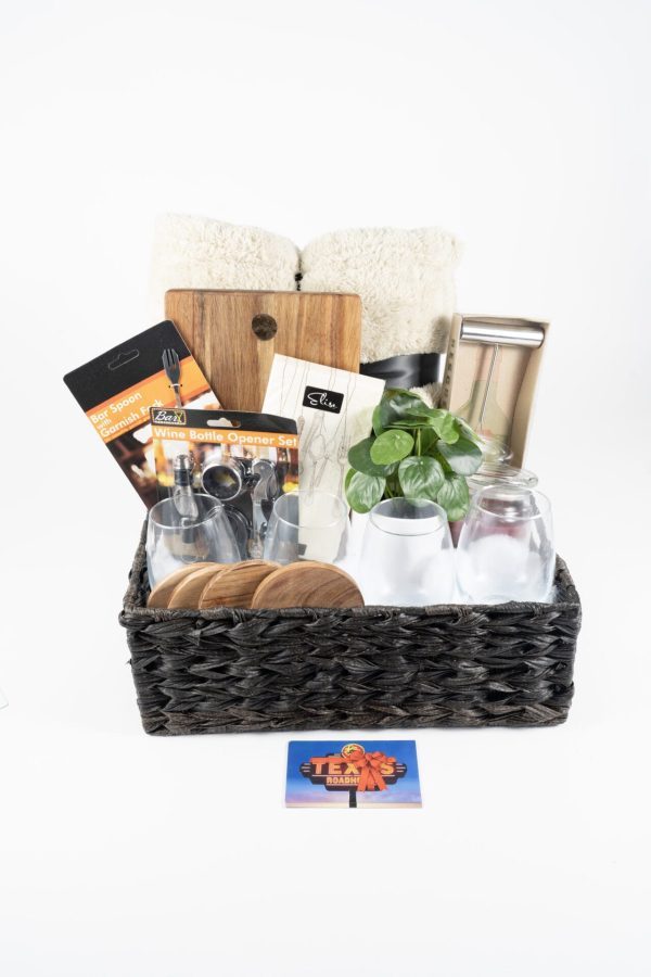 A black woven basket from our Personalized Kitchen & Home Baskets collection contains various items including a custom cutting board, four custom wine glasses, four custom coasters, $75 in Texas Roadhouse certificates, a bottle opener set, two rolled-up white towels, a small plant, and a cheese slicer. A colorful flyer with the text "Thank You" is placed in front of the basket.