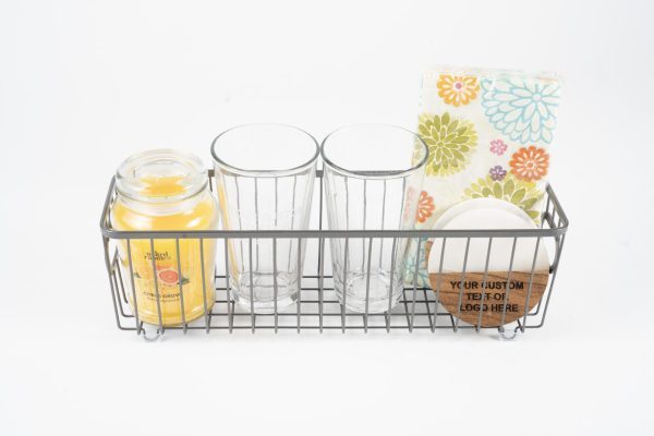 The Personalized Kitchen & Home Gift Basket with 2 Custom Glasses & 2 Custom Coasters includes a collection of items such as a yellow candle, two custom glasses, a stack of white coasters, and a colorful floral napkin set, all arranged in a metal wire basket and set against a plain white background.