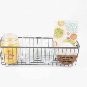 A wire basket from the Personalized Kitchen & Home Gift Basket with 2 Custom Glasses & 2 Custom Coasters collection contains a yellow scented candle, two custom glasses, a stack of colorful floral-patterned napkins, and circular custom coasters. The basket's base and sides are constructed with metal rods, offering an organized storage solution on a white background. This makes for an ideal personalized kitchen gift basket.