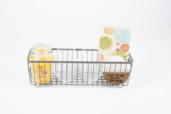 A wire basket from the Personalized Kitchen & Home Gift Basket with 2 Custom Glasses & 2 Custom Coasters collection contains a yellow scented candle, two custom glasses, a stack of colorful floral-patterned napkins, and circular custom coasters. The basket's base and sides are constructed with metal rods, offering an organized storage solution on a white background. This makes for an ideal personalized kitchen gift basket.