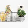 A Personalized Kitchen & Home Gift Basket, featuring a floral cloth, a glass candle jar, two custom glasses (pint, wine or juice/rocks), a stack of paper napkins, and a small potted plant with green leaves in a metal wire basket sits elegantly on a white surface against a plain white background.