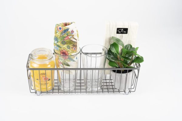 A Personalized Kitchen & Home Gift Basket, featuring a floral cloth, a glass candle jar, two custom glasses (pint, wine or juice/rocks), a stack of paper napkins, and a small potted plant with green leaves in a metal wire basket sits elegantly on a white surface against a plain white background.