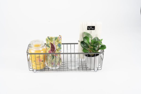 A personalized kitchen and home gift basket, featuring two custom glasses (your choice of pint, wine, or juice/rocks), contains various items including a yellow candle in a glass jar, a colorful box of tea, a small houseplant in a white pot, and a white package of napkins with a black label. This beautifully arranged basket sits against a white background—perfect as a home gift.