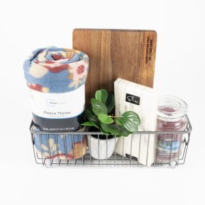 The Personalized Kitchen & Home Gift Basket with Custom Cutting Board includes a rolled-up blue floral fleece throw, a small green potted plant, a custom cutting board, a white box of tea, and a candle in a glass jar. These items are neatly arranged to create an ideal personalized kitchen gift basket.