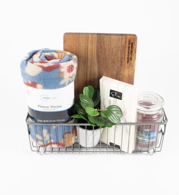 The Personalized Kitchen & Home Gift Basket with Custom Cutting Board includes a rolled-up blue floral fleece throw, a small green potted plant, a custom cutting board, a white box of tea, and a candle in a glass jar. These items are neatly arranged to create an ideal personalized kitchen gift basket.