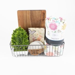 The Personalized Kitchen & Home Gift Basket with 2 Custom Coasters and a Cutting Board includes a charming array of items in a stylish metal wire container, featuring a small cutting board, an artificial green potted plant, a floral-patterned fleece throw, a floral box, a white candle, and a small rectangular sign with text. Each item is meticulously arranged against a plain white background for an elegant presentation.