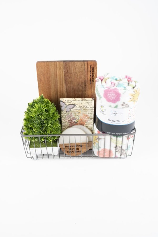 The Personalized Kitchen & Home Gift Basket with 2 Custom Coasters and a Cutting Board includes a charming array of items in a stylish metal wire container, featuring a small cutting board, an artificial green potted plant, a floral-patterned fleece throw, a floral box, a white candle, and a small rectangular sign with text. Each item is meticulously arranged against a plain white background for an elegant presentation.