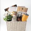 The Adult Gambling Night Basket includes a small potted plant, wooden cutting boards, a soft throw blanket, a yellow travel mug, a box with small compartments, a decorative tile, and an adult-themed cloth towel adorned with fruit designs. The basket is showcased against a plain white background.