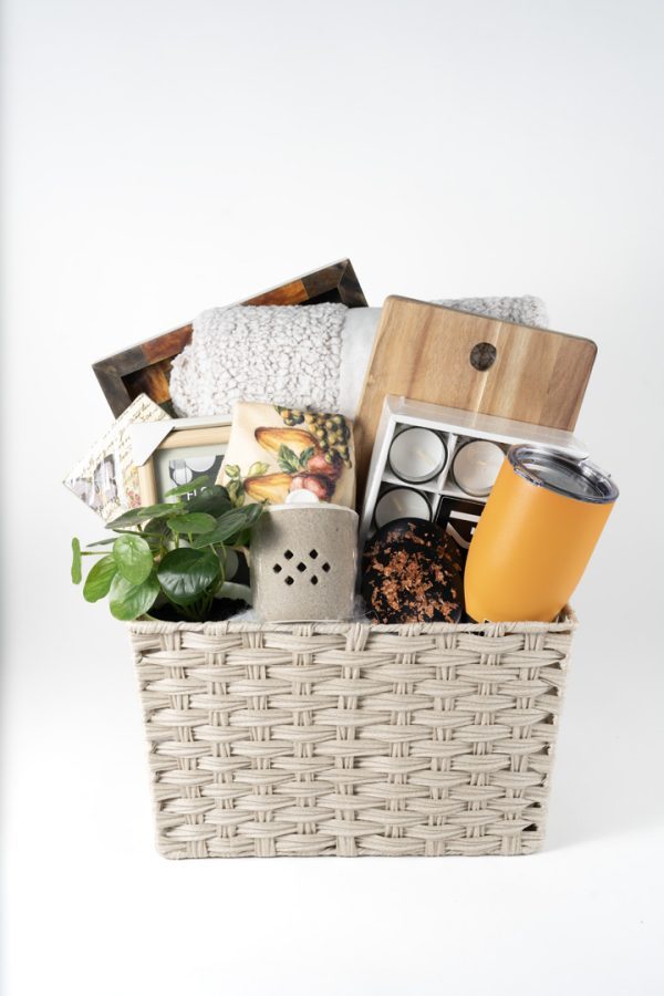 The Adult Gambling Night Basket includes a small potted plant, wooden cutting boards, a soft throw blanket, a yellow travel mug, a box with small compartments, a decorative tile, and an adult-themed cloth towel adorned with fruit designs. The basket is showcased against a plain white background.