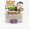 The Large Kitchen & Home Basket with $75 in Texas Roadhouse Certificates is tastefully filled with charming items including a wooden cutting board, a blue towel, a floral-themed towel, a small potted plant, and candles. The beige woven home basket is elegantly presented against a plain white background.