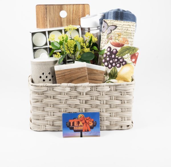 The Large Kitchen & Home Basket with $75 in Texas Roadhouse Certificates is tastefully filled with charming items including a wooden cutting board, a blue towel, a floral-themed towel, a small potted plant, and candles. The beige woven home basket is elegantly presented against a plain white background.