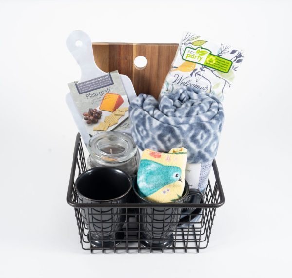 The Kitchen & Home Basket with Coffee Mugs includes two black ceramic coffee mugs, a rolled-up grey patterned blanket, a transparent jar with a lid, a rectangular cutting board for the kitchen, colorful socks with bird designs, eco-friendly party napkins, and a cheese and cracker set.