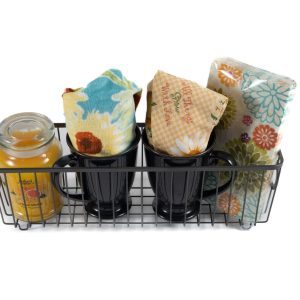 The "Kitchen & Home - Small Gift Basket with Mugs" features a metal wire basket that holds a yellow scented candle, two black mugs each with a floral-printed cloth inside, and two additional floral-printed napkins or cloths neatly stacked at the end. The items are beautifully arranged to create a vibrant and colorful gift basket perfect for any kitchen or home setting.