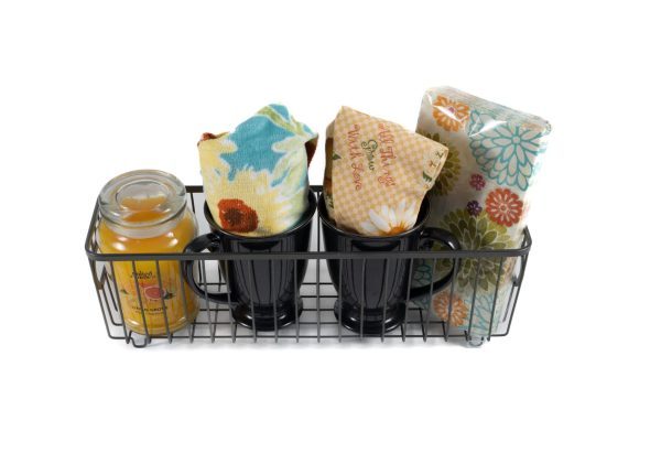 The "Kitchen & Home - Small Gift Basket with Mugs" features a metal wire basket that holds a yellow scented candle, two black mugs each with a floral-printed cloth inside, and two additional floral-printed napkins or cloths neatly stacked at the end. The items are beautifully arranged to create a vibrant and colorful gift basket perfect for any kitchen or home setting.