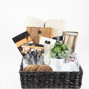 The Personalized Kitchen & Home Gift Basket with 4 Custom Glasses, 4 Custom Coasters, and a Cutting Board contains various household items, including rolled white towels, a small plant, glass jars, a metal tumbler, Bar Spoon and Wine Bottle Opener Sets, and other kitchen essentials against a white background.