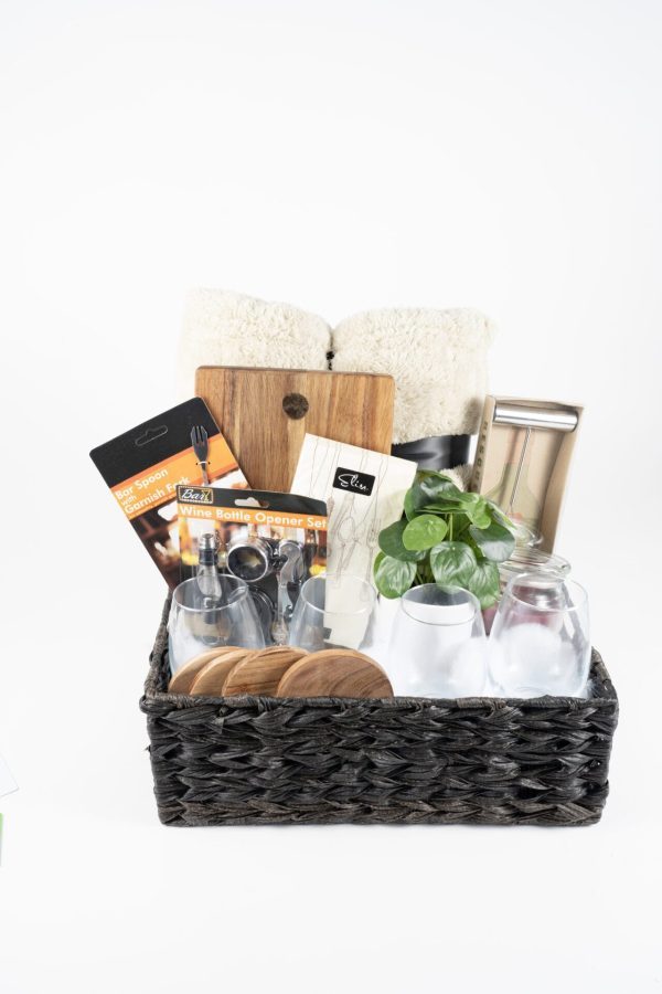 The Personalized Kitchen & Home Gift Basket with 4 Custom Glasses, 4 Custom Coasters, and a Cutting Board contains various household items, including rolled white towels, a small plant, glass jars, a metal tumbler, Bar Spoon and Wine Bottle Opener Sets, and other kitchen essentials against a white background.