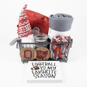 A Buckeye Gift Basket - Medium includes a variety of Ohio State-themed items such as a plush gnome, a red and gray blanket, jar candles, coasters, an oven mitt, and a rolled-up banner. In front of the basket is a sign that reads "FOOTBALL IS MY FAVORITE SEASON.