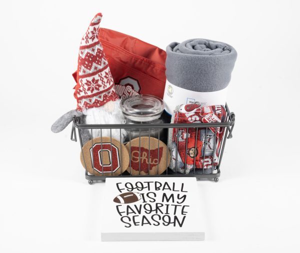 A Buckeye Gift Basket - Medium includes a variety of Ohio State-themed items such as a plush gnome, a red and gray blanket, jar candles, coasters, an oven mitt, and a rolled-up banner. In front of the basket is a sign that reads "FOOTBALL IS MY FAVORITE SEASON.