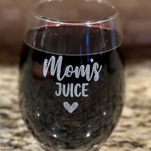 A stemless Mom's Juice Wine Glass filled with a dark red liquid, likely wine, stands on a granite countertop. The front of the wine glass displays the text "Mom's Juice" with a small heart symbol below it.