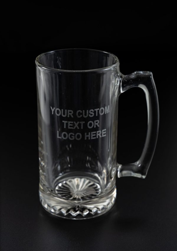 The Glass Mug - Single is a clear glass mug featuring an inscription on the front that reads "YOUR CUSTOM TEXT OR LOGO HERE." This single glass mug comes with a simple, sturdy handle and a patterned base, set against a dark background.