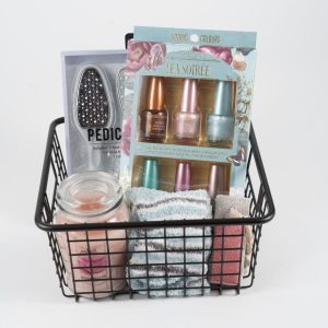 The Manicure/Pedicure Basket comprises a black wire basket filled with a variety of self-care items, including a pedicure tool, a set of four pastel nail polishes for manicures, a peach-scented candle, and several folded washcloths in assorted colors.