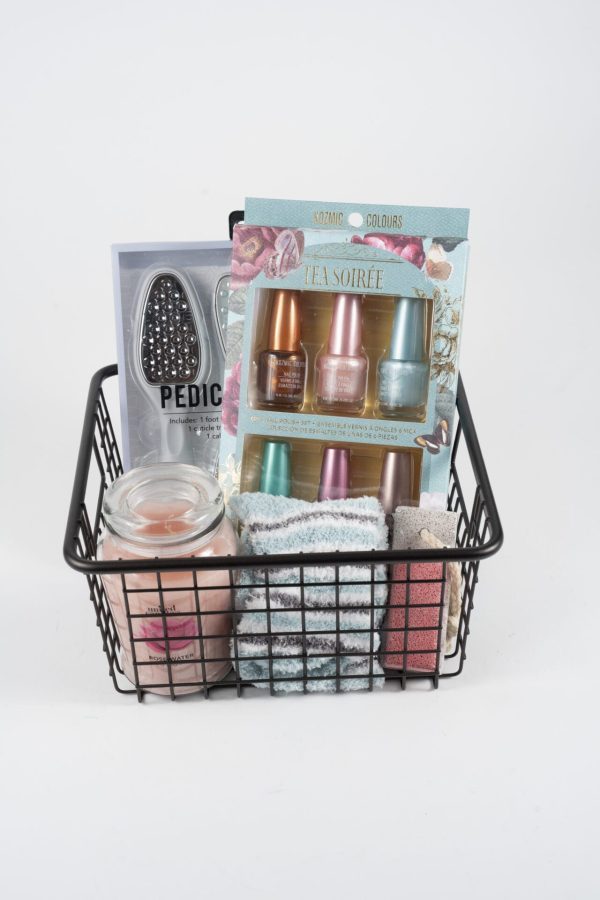 The Manicure/Pedicure Basket comprises a black wire basket filled with a variety of self-care items, including a pedicure tool, a set of four pastel nail polishes for manicures, a peach-scented candle, and several folded washcloths in assorted colors.